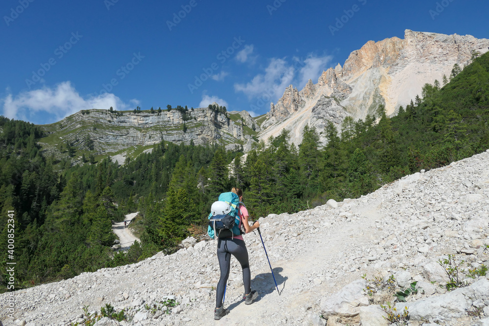 A woman with a hiking backpack hiking on a gravelled road in high Italian Dolomites. There is a dence forest at the foothill, and steep and sharp mountain chain in the back. Discovering and exploring