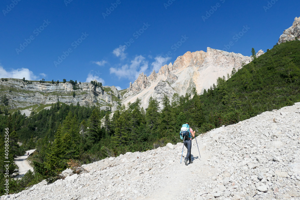A woman with a hiking backpack hiking on a gravelled road in high Italian Dolomites. There is a dence forest at the foothill, and steep and sharp mountain chain in the back. Discovering and exploring