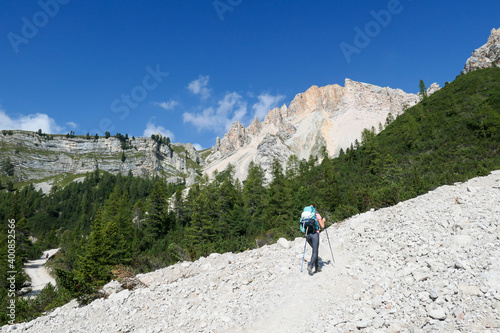 A woman with a hiking backpack hiking on a gravelled road in high Italian Dolomites. There is a dence forest at the foothill, and steep and sharp mountain chain in the back. Discovering and exploring