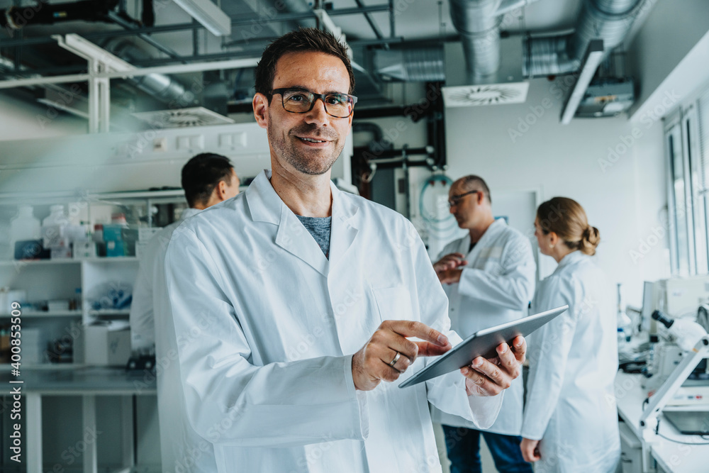 Smiling male scientist using digital tablet while standing with coworker in background at laboratory Smiling male scientist using digital tablet while standing with coworker in background at laboratory