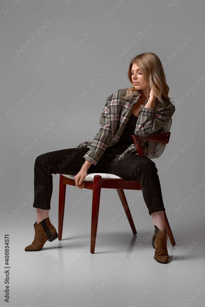 Fototapeta premium beautiful blonde woman with curly hair in jacket sitting on chair on studio background.