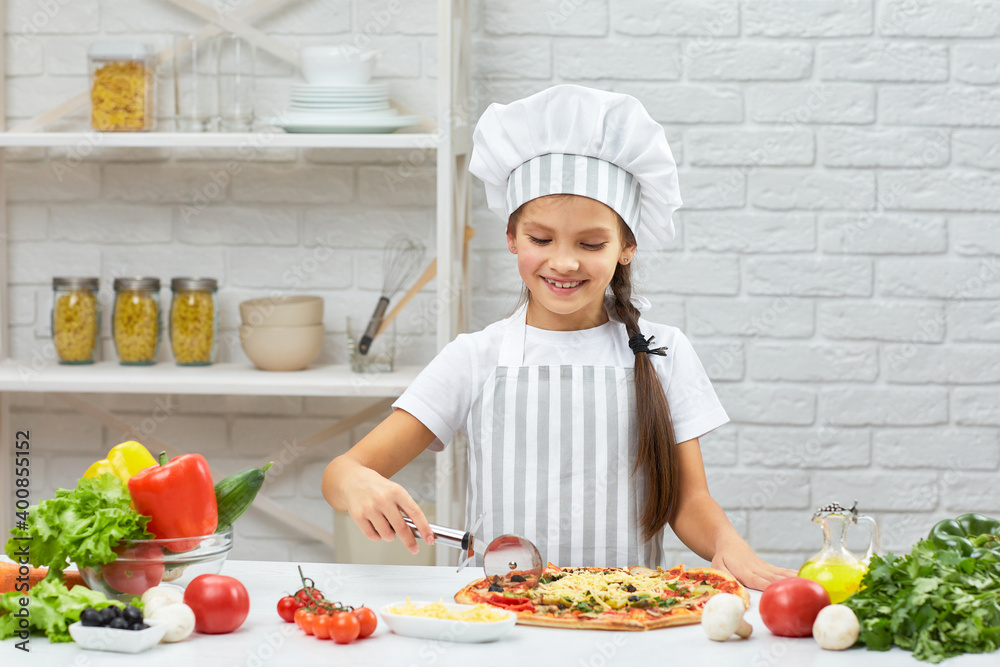 cute smiling little girl in chef hat and an apron cutting pizza at kitchen cute smiling little girl in chef hat and an apron cutting pizza at kitchen
