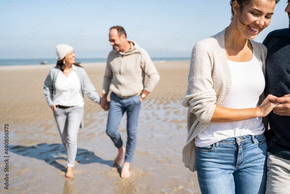 Smiling woman holding man hand while walking with couple in background at beach Smiling woman holding man hand while walking with couple in background at beach