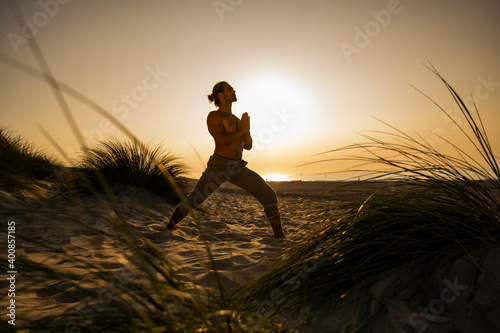 Shirtless young man practicing warrior position yoga with hands clasped at beach against clear sky during sunset photo