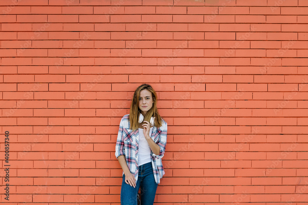Young woman with headphone around her neck standing against brick wall
