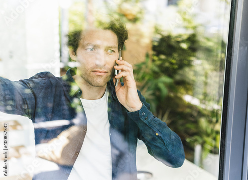 Mid adult man talking on smart phone while looking through window at home