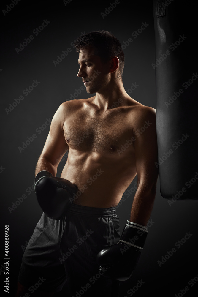 Naklejka premium Young handsome male boxer in black gloves standing near punching bag on dark background