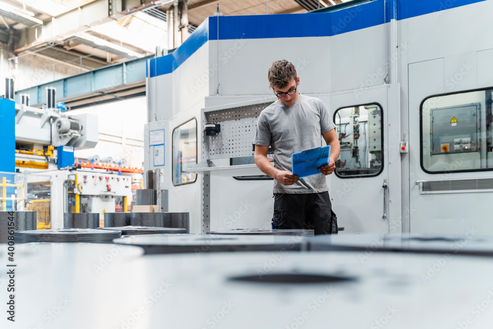 Male engineer holding clipboard while standing by machinery in illuminated factory Male engineer holding clipboard while standing by machinery in illuminated factory