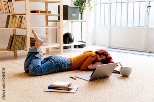 Redhead woman with book and laptop lying on carpet at home photo