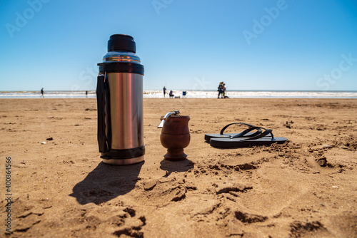 A thermos with a mate and a pair of flip flops resting on the sand on the beach with people walking in the background