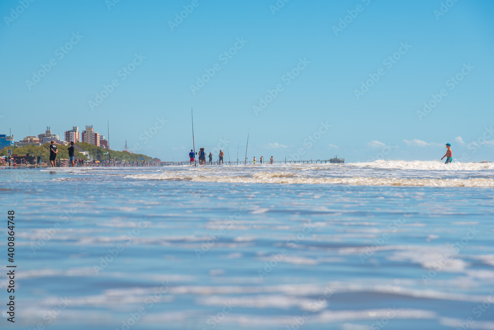 Mar del Tuyú, Buenos Aires, Argentina; December 06, 2020: View of the beach with a lot of fishermen and a pier in the background from inside the water Mar del Tuyú, Buenos Aires, Argentina; December 06, 2020: View of the beach with a lot of fishermen and a pier in the background from inside the water