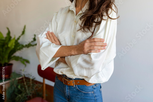 Woman wearing white shirt with arms crossed standing against wall at home