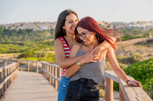 Cheerful teenage girls embracing on boardwalk at beach against clear sky