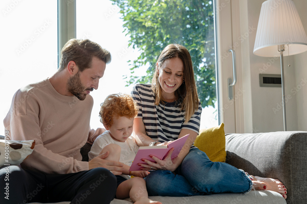 Smiling woman reading picture book while sitting by son and man on sofa in living room at home Smiling woman reading picture book while sitting by son and man on sofa in living room at home