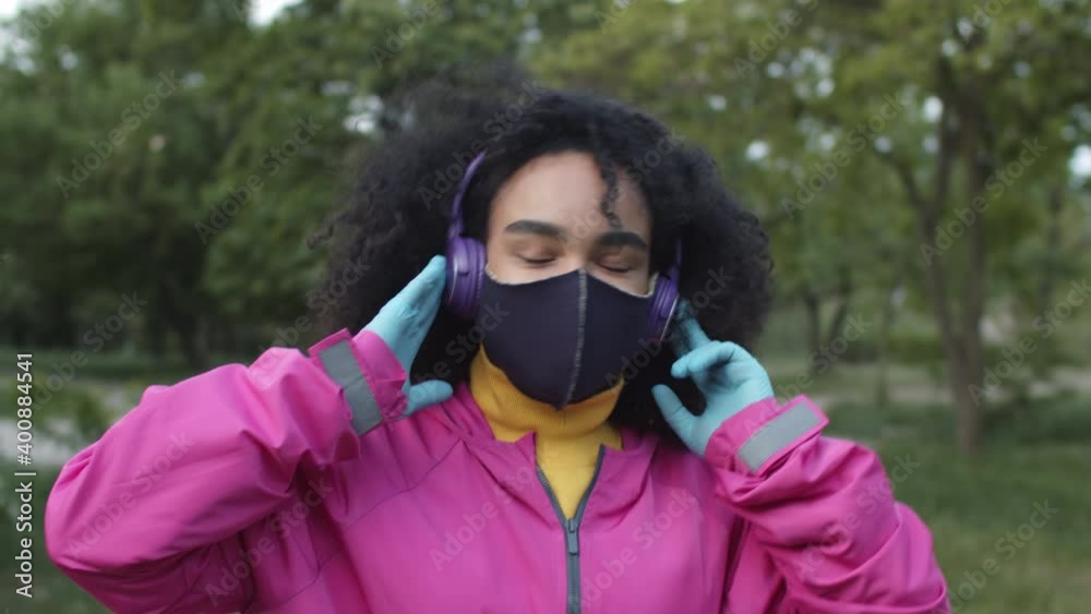 Beautiful african american woman walking in park in spring, wearing covid-19 face mask and gloves, listening music in headphones and looking happy Beautiful african american woman walking in park in spring, wearing covid-19 face mask and gloves, listening music in headphones and looking happy