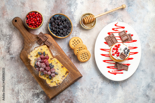 top view raisin cake slices with raspberries on cutting board bowl with cornels ans raisins biscuits honeycomp stick in honey bowl and cake with chocolate on white plate on marble ground photo