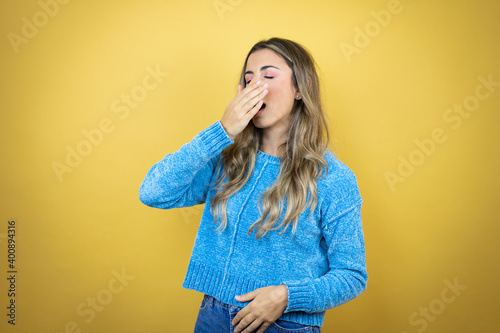 Pretty blonde woman with long hair standing over yellow background bored yawning tired covering mouth with hand. Restless and sleepiness.
