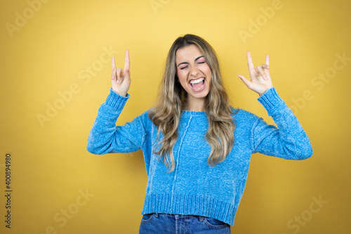 Pretty blonde woman with long hair standing over yellow background shouting with crazy expression doing rock symbol with hands up