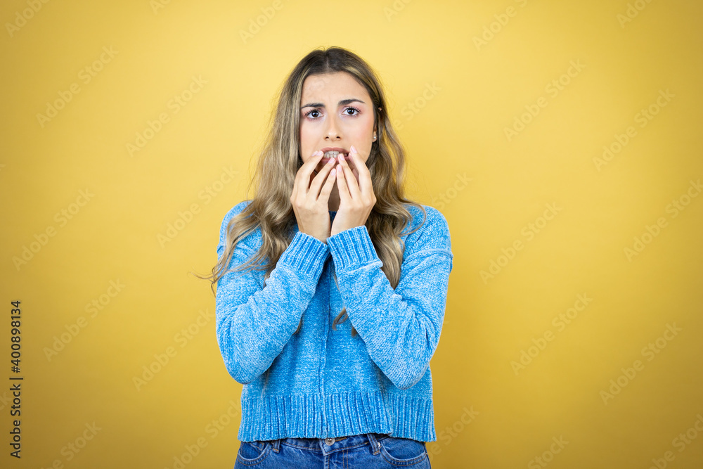 Pretty blonde woman with long hair standing over yellow background nervous, biting her nails