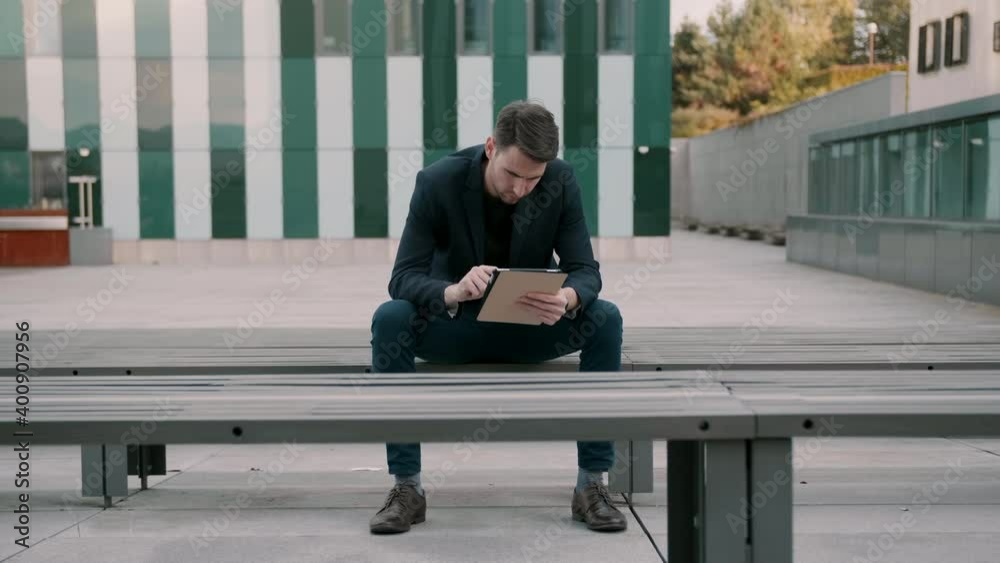 Businessman sitting on a bench and being focused while typing on his tablet Businessman sitting on a bench and being focused while typing on his tablet