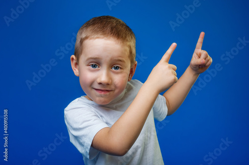 Close-up portrait of a surprised cheerful child pointing two fingers to the side on a blue background. A copy of the space.