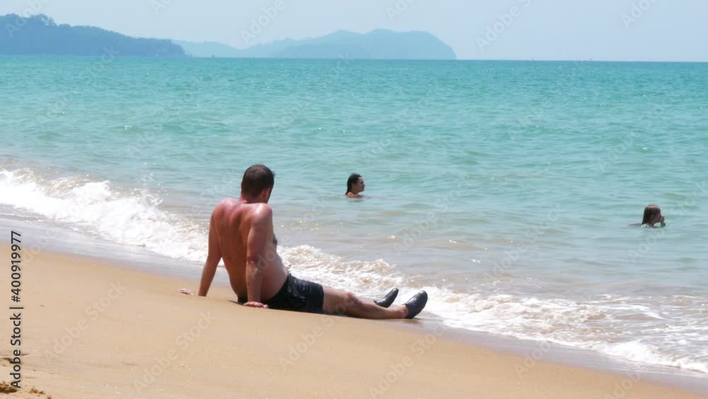 Man sits on sandy tropical beach watching family swimming in the sea on vacation. Static shot. Man sits on sandy tropical beach watching family swimming in the sea on vacation. Static shot.