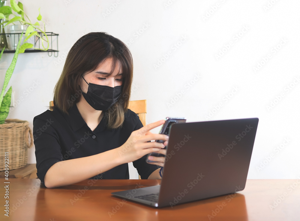 Asian woman wearing black protective face mask, sitting at wooden table with computer laptop using mobile phone. white wall with house plant background.