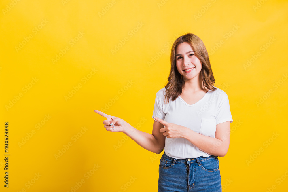 Asian Thai happy portrait beautiful cute young woman standing wear white t-shirt pointing finger away side looking to camera, studio shot isolated on yellow background with copy space Asian Thai happy portrait beautiful cute young woman standing wear white t-shirt pointing finger away side looking to camera, studio shot isolated on yellow background with copy space