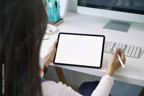 Backside view of woman working with mockup blank white screen tablet at working desk. White screen tablet for graphic montage.