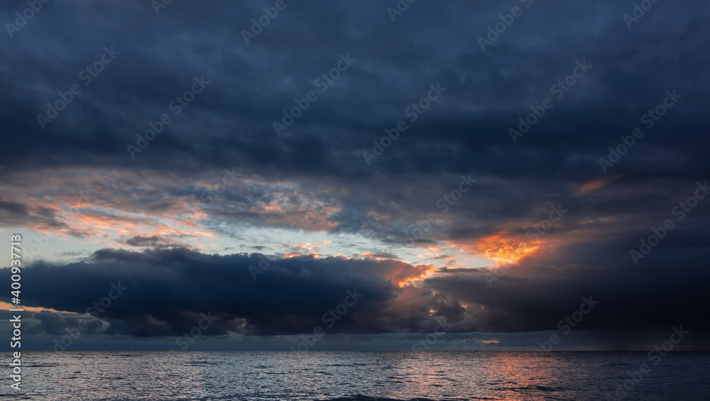 The sky is almost entirely covered with dark dense clouds. In their break is a bright orange highlight from the sun's rays. Reflection on the shiny surface of the sea. It's raining in the distance. The sky is almost entirely covered with dark dense clouds. In their break is a bright orange highlight from the sun's rays. Reflection on the shiny surface of the sea. It's raining in the distance.