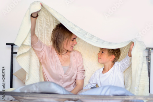 mom and child have fun in the bedroom on the bed, play under the blanket. Young mother with her little son dressed playing in the bed at the weekend together, lazy morning, warm and cozy scene.