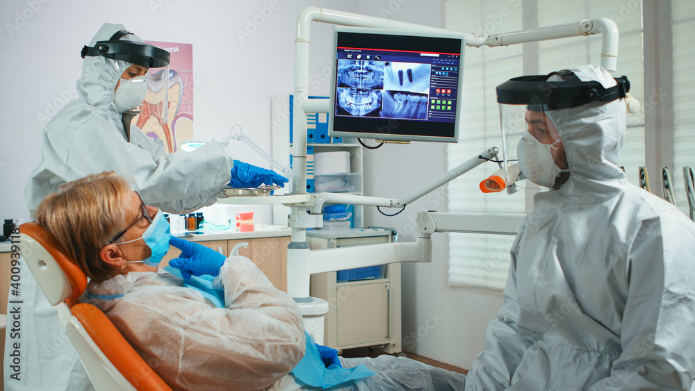 Dentist doctor with face shield pointing on stomatological monitor explaining dental x-ray to patient during global pandemic. Nurse and orthodontist wearing protection suit, coverall, mask and gloves Dentist doctor with face shield pointing on stomatological monitor explaining dental x-ray to patient during global pandemic. Nurse and orthodontist wearing protection suit, coverall, mask and gloves