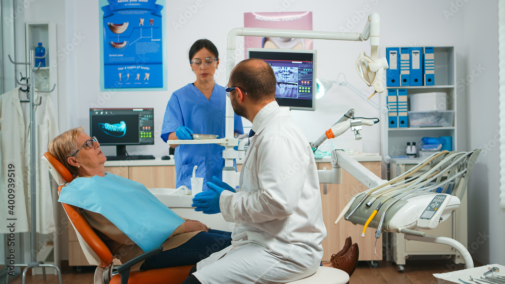 Obraz premium Dentistry doctor explaining to old patient the dental process of intervention. Orthodontist speaking to woman with toothache sitting on stomatological chair while nurse preparing tools for examination