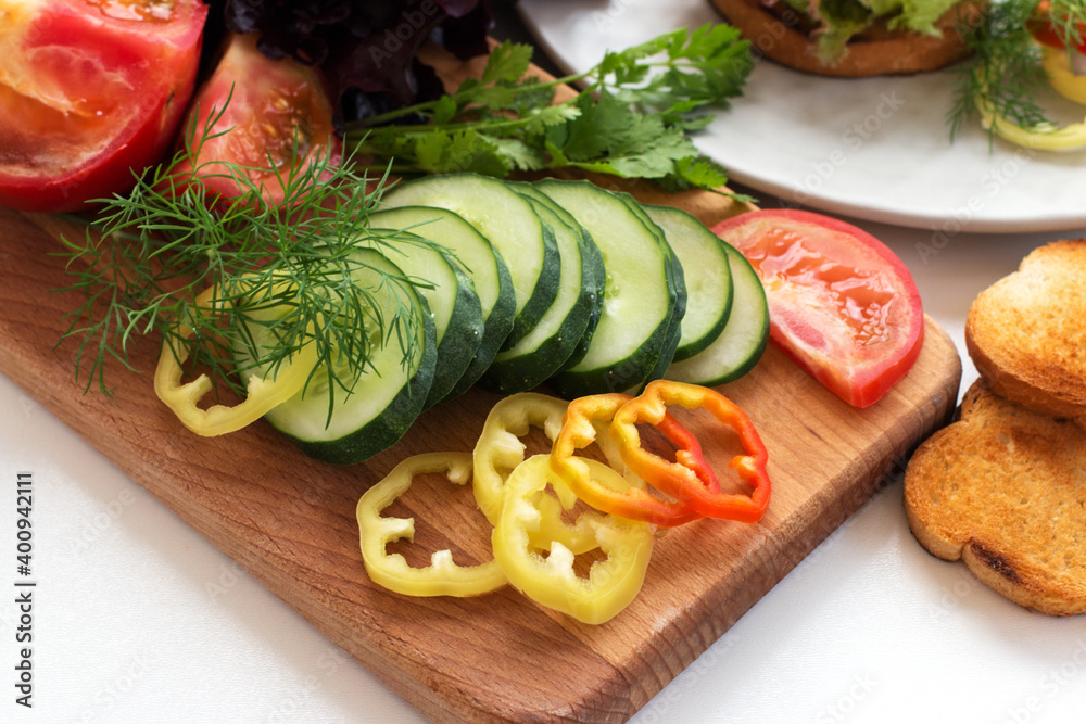 Sliced ​​pieces of green cucumbers, red tomatoes, yellow peppers, decorated with fresh dill and parsley, lie on a wooden board. There are croutons nearby. Diet. Cooking. Close-up.