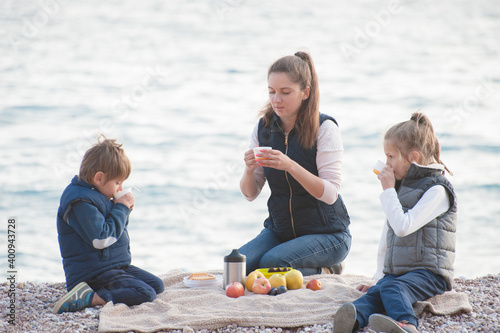 active caucasian sport family in sleeveless jackets sitting on sea beach during spring picnic drinking hot tea