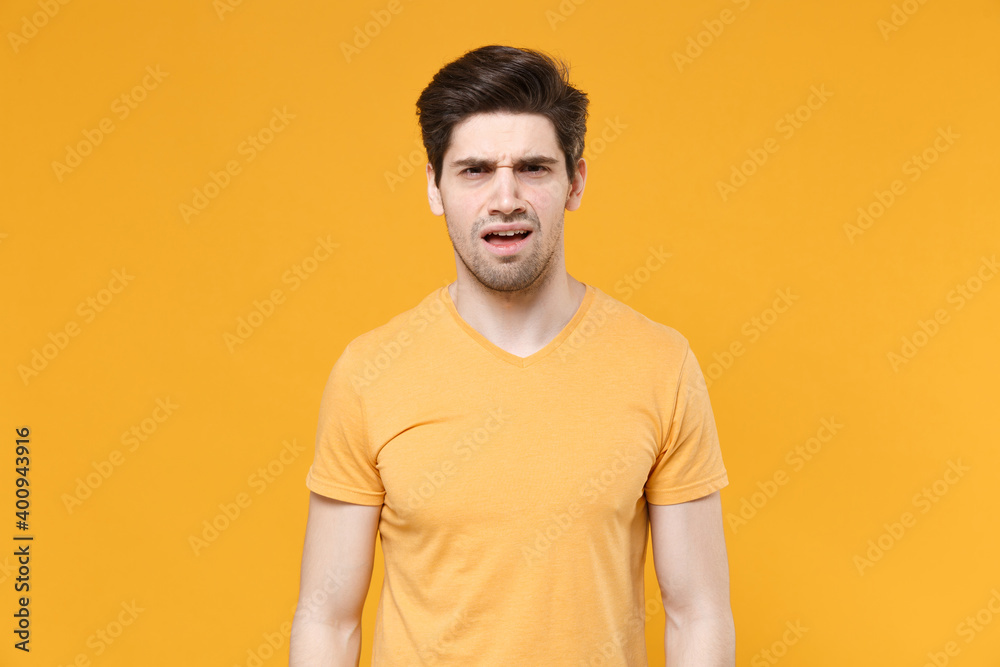 Young disgusted confused displeased unshaved caucasian handsome man 20s years old wearing casual basic blank print design t-shirt looking camera isolated on yellow color background studio portrait.