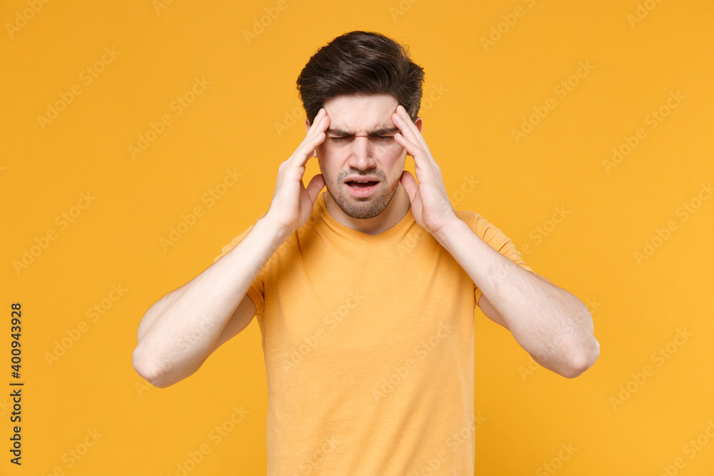 Young sick ill unshaved caucasian man 20s in basic blank print design t-shirt put hands on head rub temples having headache migraine, feel bad seedy isolated on yellow background studio portrait. Young sick ill unshaved caucasian man 20s in basic blank print design t-shirt put hands on head rub temples having headache migraine, feel bad seedy isolated on yellow background studio portrait.