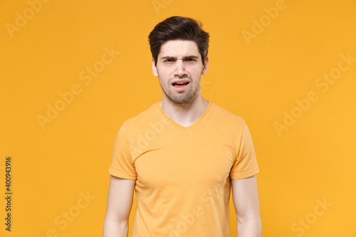 Young disgusted confused displeased unshaved caucasian handsome man 20s years old wearing casual basic blank print design t-shirt looking camera isolated on yellow color background studio portrait. © ViDi Studio