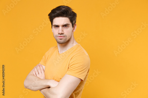 Young serioius unshaved caucasian handsome man 20s wearing casual basic blank print design t-shirt holding crossed hands folded looking camera isolated on yellow color background studio portrait.