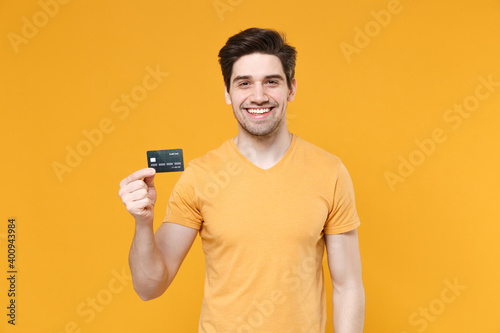 Young smiling cheerful unshaved successful caucasian happy man 20s wearing casual basic blank print design t-shirt hold in hand credit bank card isolated on yellow color background studio portrait.