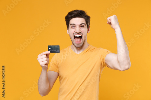 Young happy unshaved successful man in basic blank print t-shirt hold in hand credit bank card winner gesture with clenching fists saying yes isolated on yellow color background studio portrait.