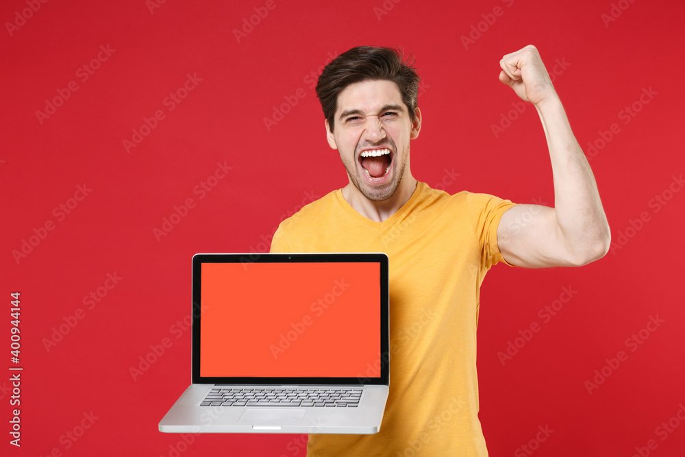 Young happy unshaved happy man in yellow t-shirt using laptop pc computer blank screen workspace area copy space mock up doing winner gesture clenching fist isolated on red background studio portrait