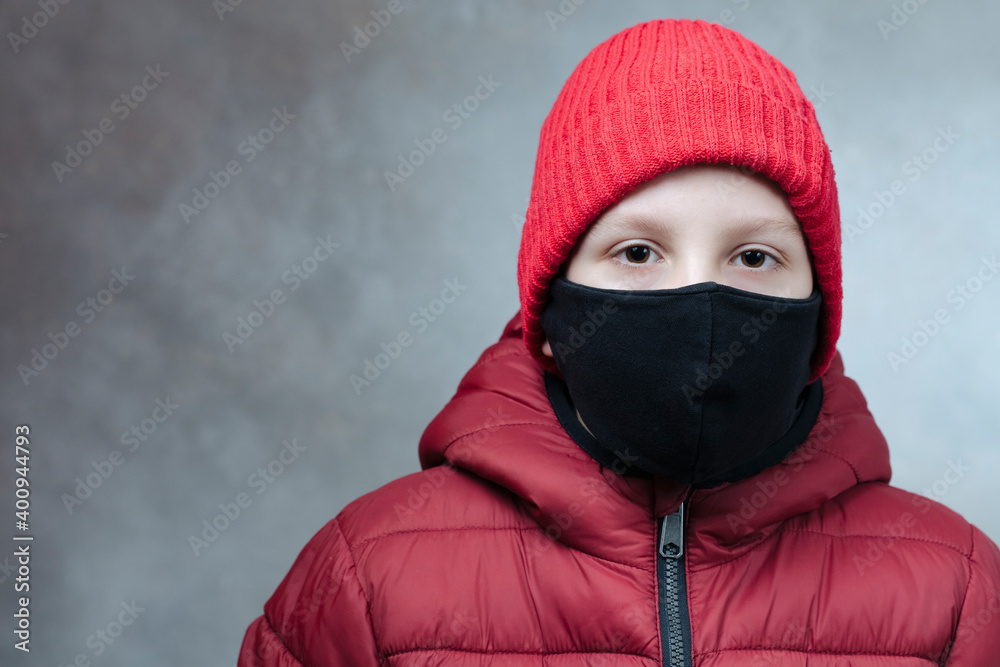 Boy with safety mask from coronavirus in winter jacket and red  hat on damage wall. Covid-19 concept