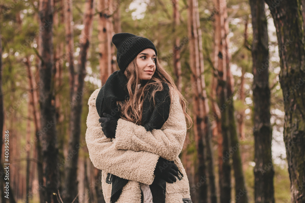 woman of model appearance dressed in winter clothes, black hat, gloves and a scarf posing while in a pine forest woman of model appearance dressed in winter clothes, black hat, gloves and a scarf posing while in a pine forest