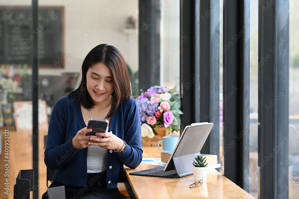 Fototapeta premium Cheerful female chatting online in social networks with friend on smart phone while sitting in office.