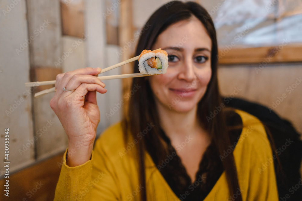 Close-up attractive woman portrait at the restaurant showing sushi roll in chopsticks looking at the camera.
