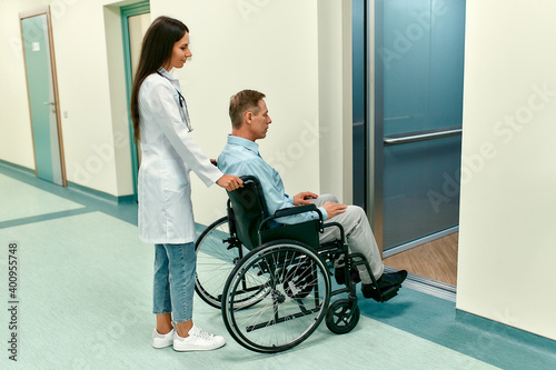 Young beautiful woman doctor with her disabled elderly patient in a wheelchair approached the elevator of a modern hospital.