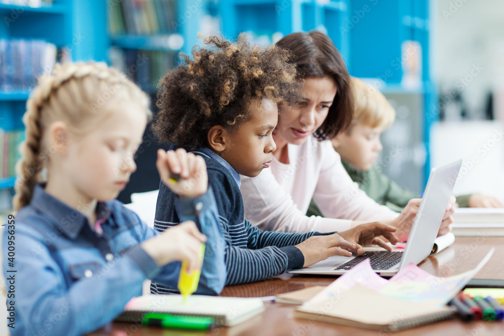 Female teacher explaining task on laptop computer screen to mixed race elementary student while his classmates studying sitting at desk nearby in school classroom, side view Female teacher explaining task on laptop computer screen to mixed race elementary student while his classmates studying sitting at desk nearby in school classroom, side view