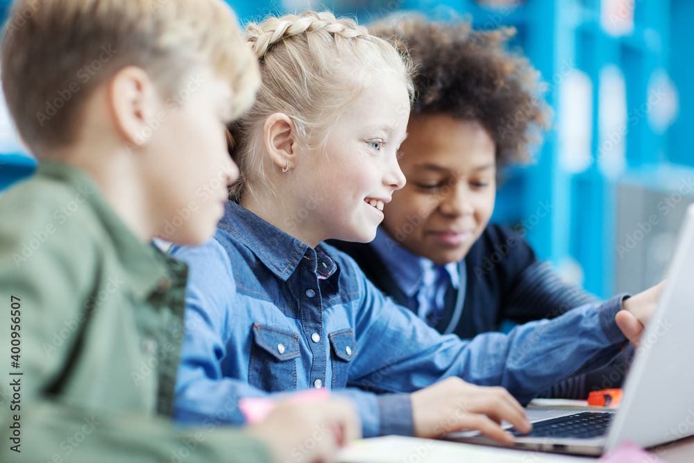 © Comeback Images - Group of three elementary students sitting at desk in classroom and studying or playing on laptop; focus on smiling blonde girl pointing at screen