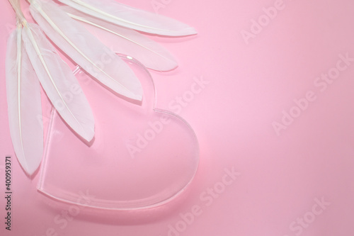 Transparent glass heart and white feathers on pale pink background. Place for your text. Valentine card, declaration of love, postcard, wedding invitation.